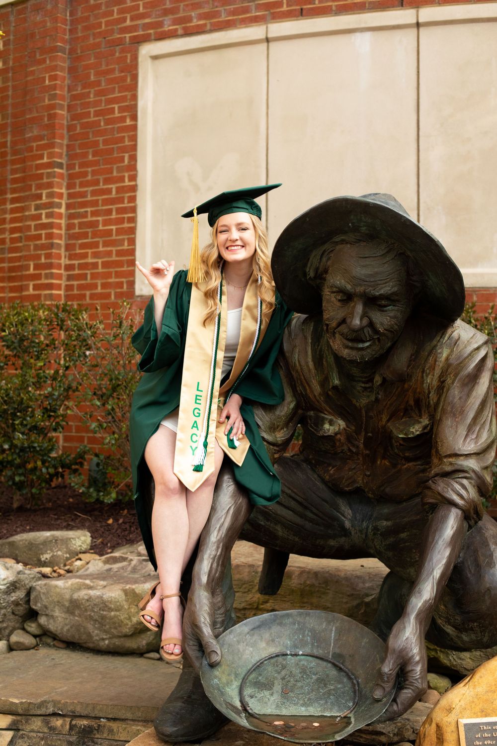 Graduate poses playfully next to bronze statue on university campus.