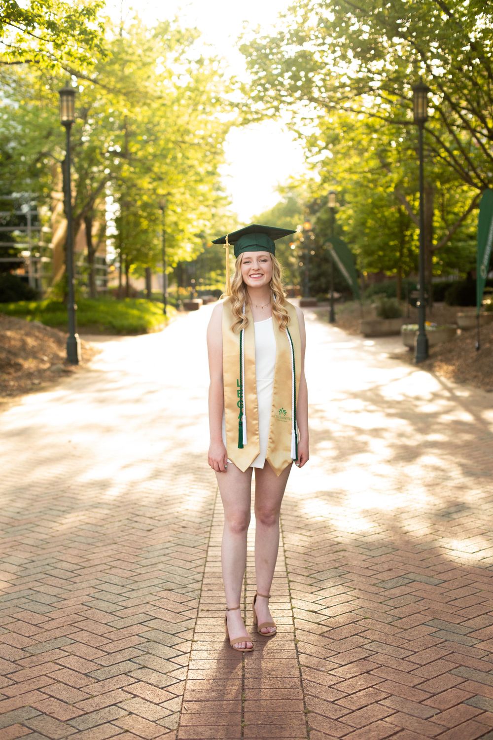 Graduate in green regalia stands on sunlit brick pathway surrounded by trees.