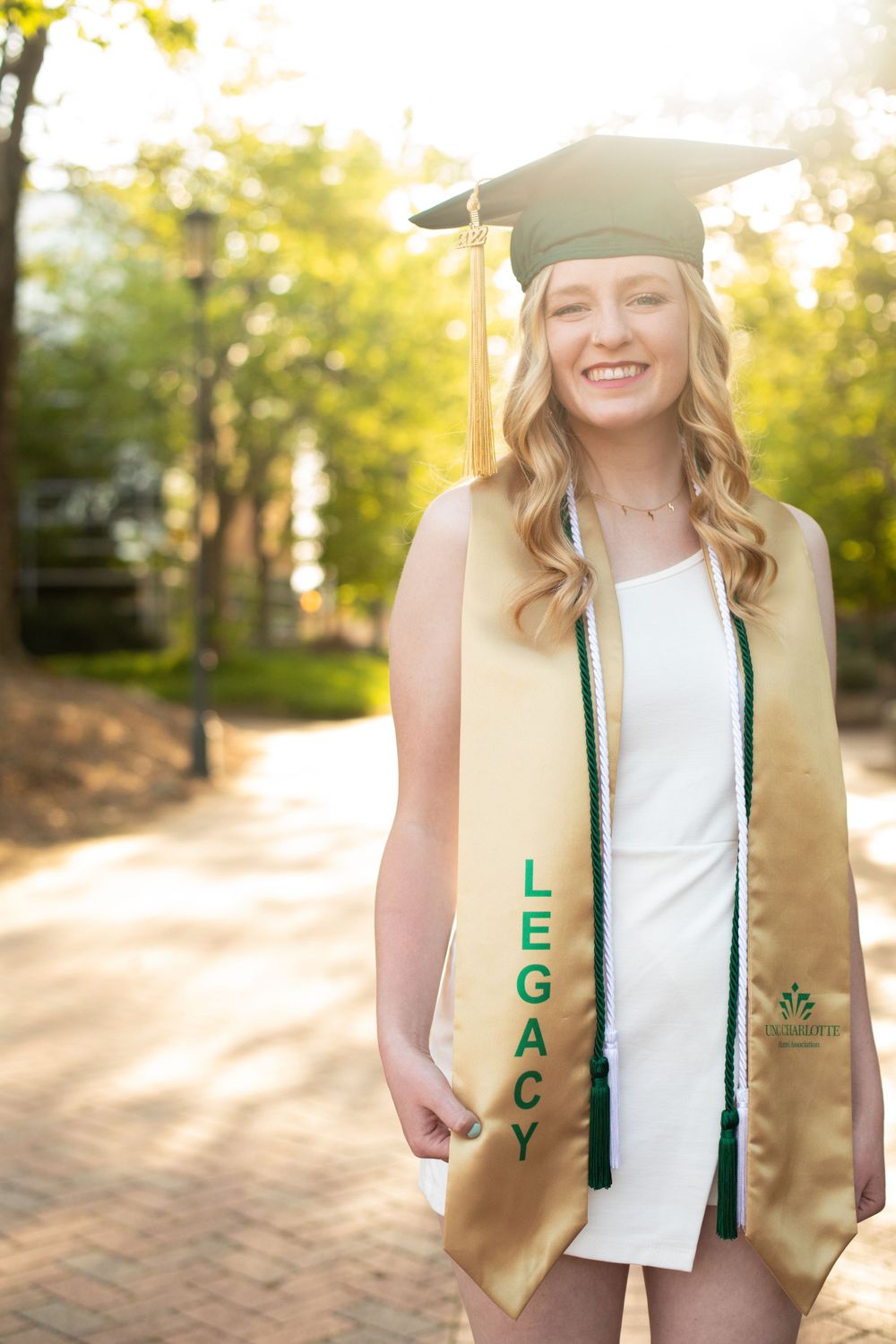 Graduate in cap and gold stole poses on sunny campus pathway.