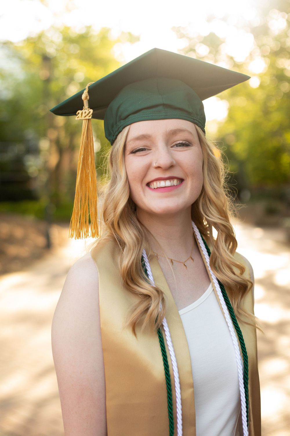 Graduate smiles radiantly in green cap with gold tassel during outdoor portrait session.