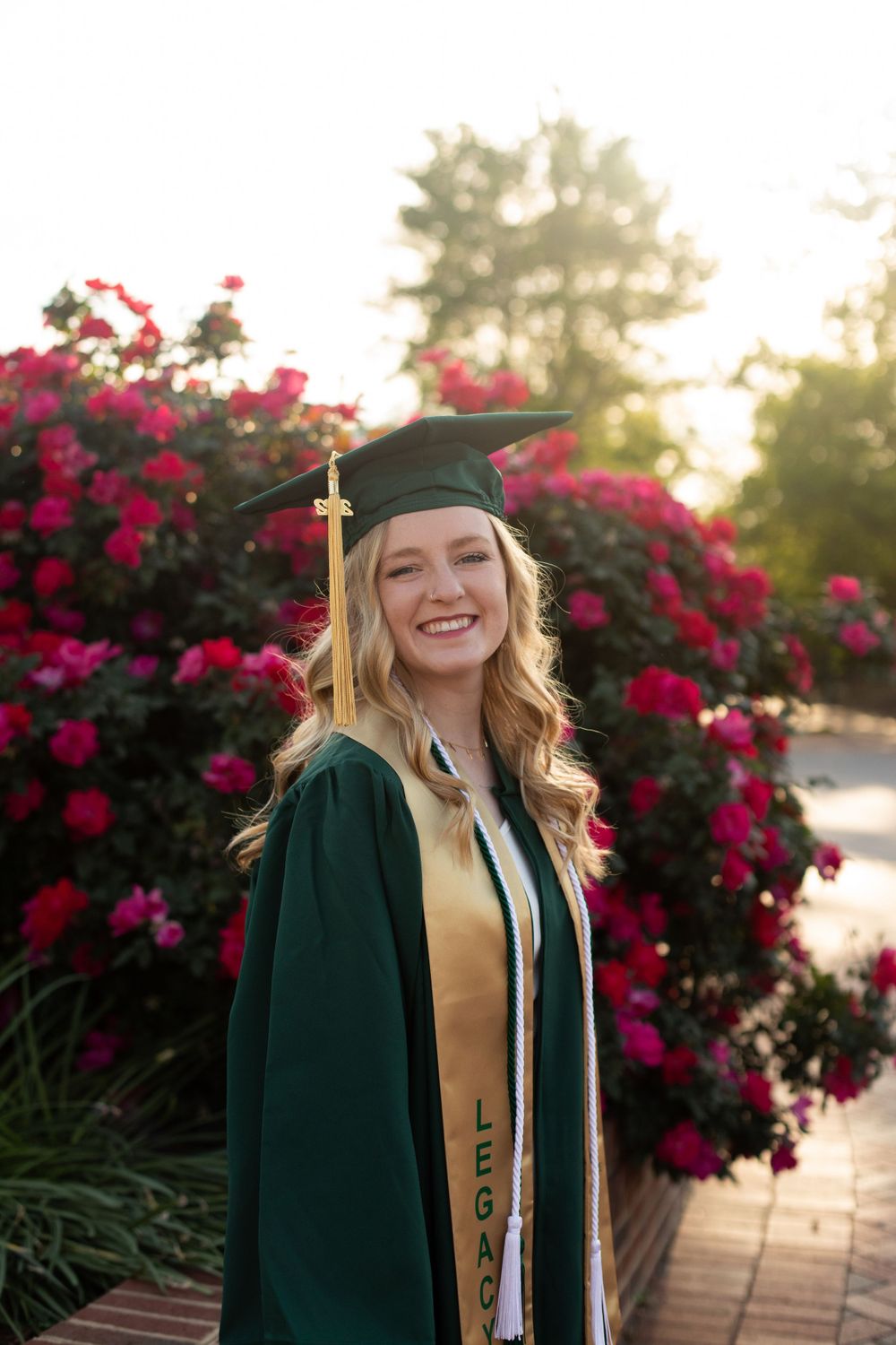 Graduate in green cap and gown poses against vibrant red flowers during golden hour sunset.