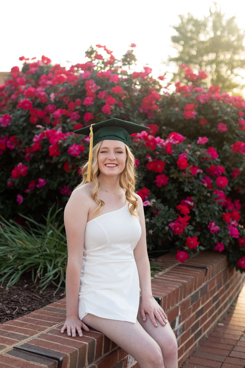 Graduate in white dress and cap sits on brick ledge with blooming red flowers in background.