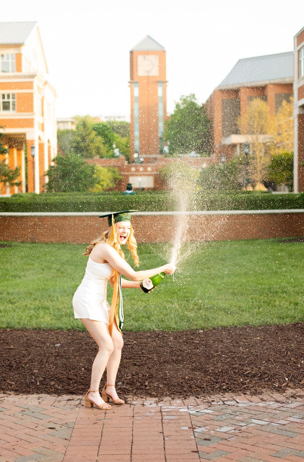 Graduate in white dress pops champagne on campus with clock tower in background.