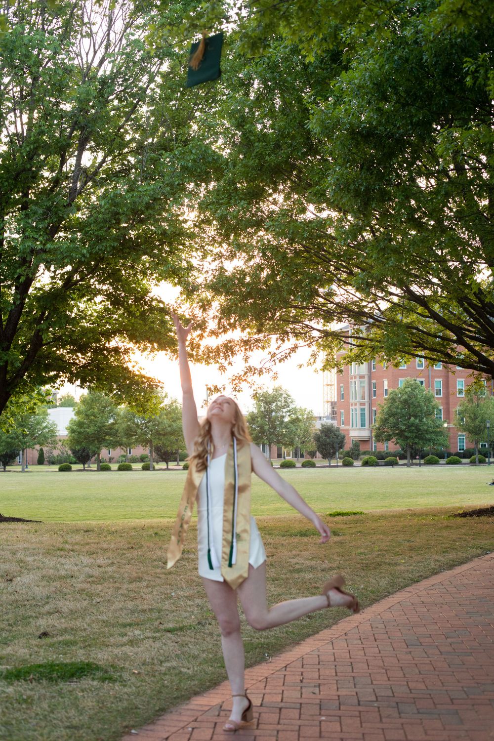 Graduate in white dress jumps joyfully on brick pathway with campus buildings in background.