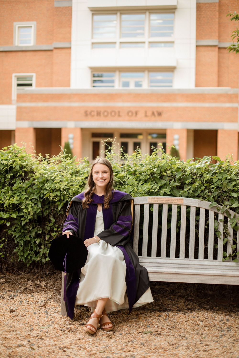 Graduate in academic regalia sitting on bench outside School of Law building.