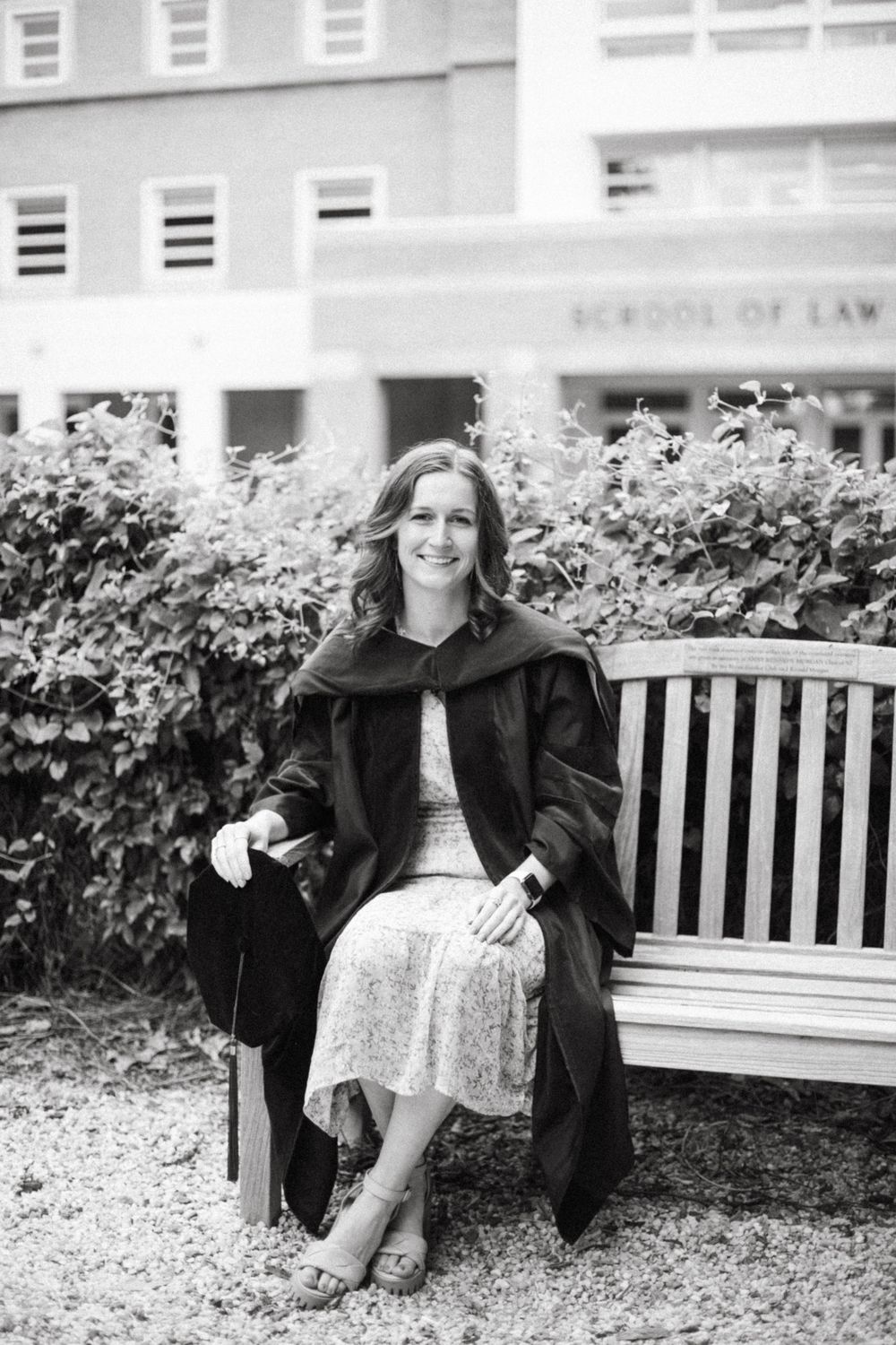 Graduate in cap and gown sitting on bench outside law school building.