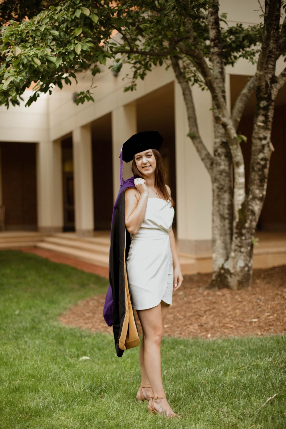 Graduate in white dress and academic gown posing on grass near campus trees.