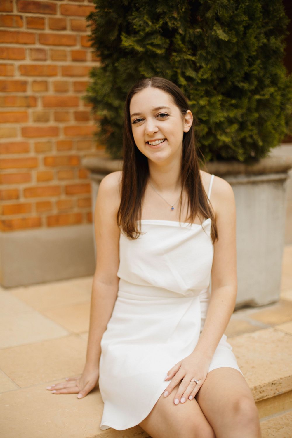 Graduate in white dress sitting casually outside brick building.