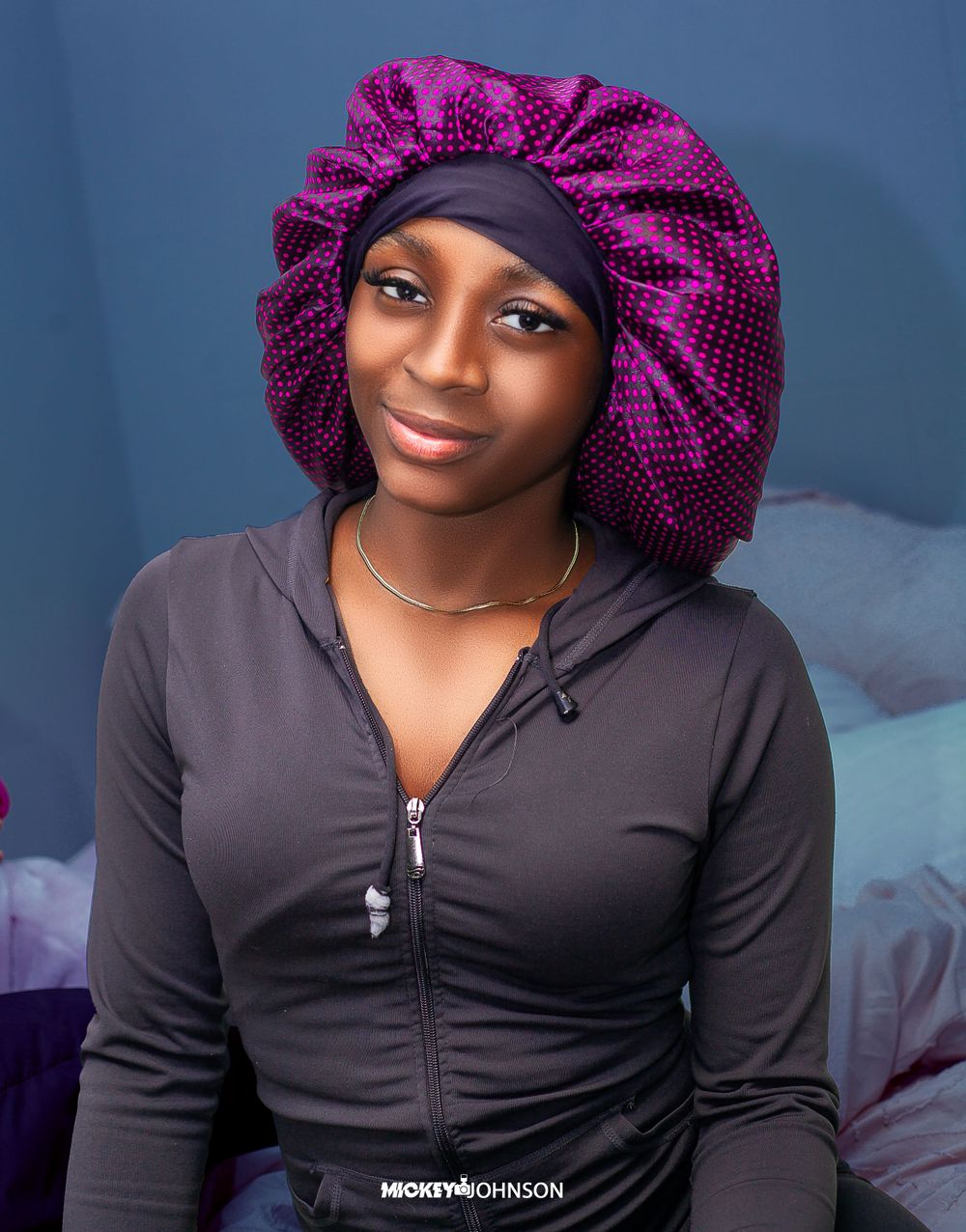 Portrait of someone wearing a purple bonnet and black zip-up hoodie against a blue background.