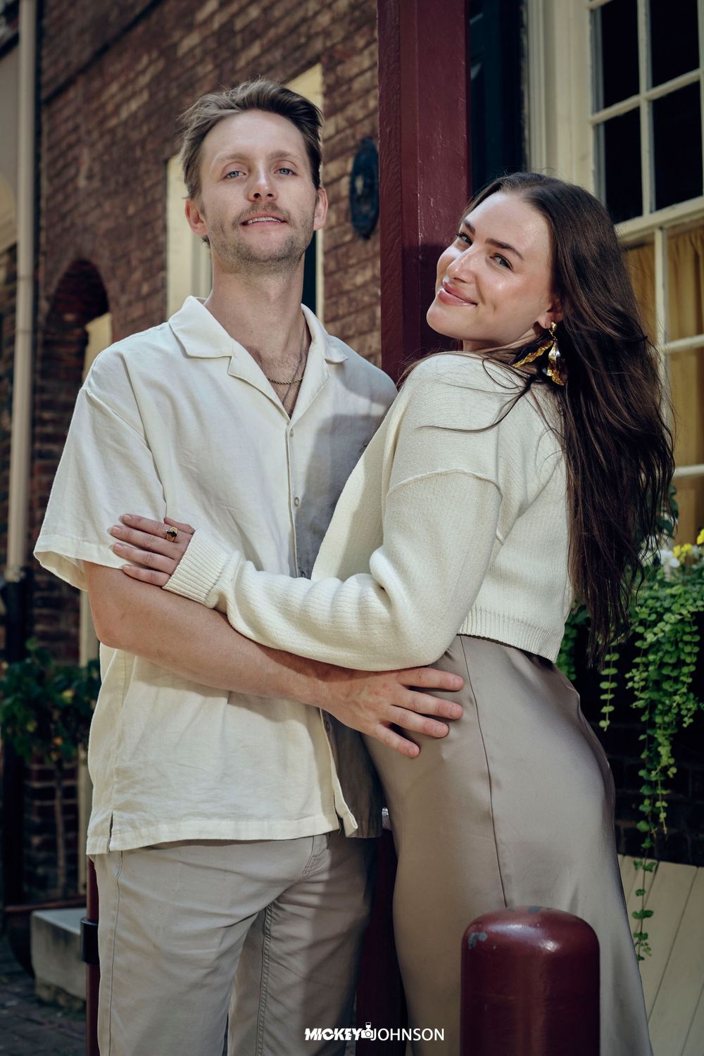 Couple in matching cream outfits embracing against brick building exterior.
