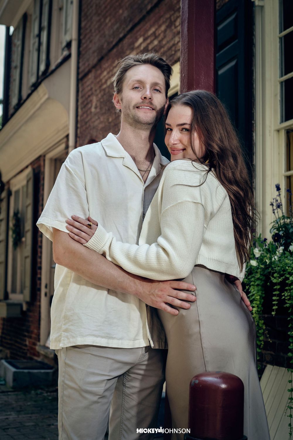 A couple embraces while wearing matching cream-colored outfits in front of a brick building on a city street.