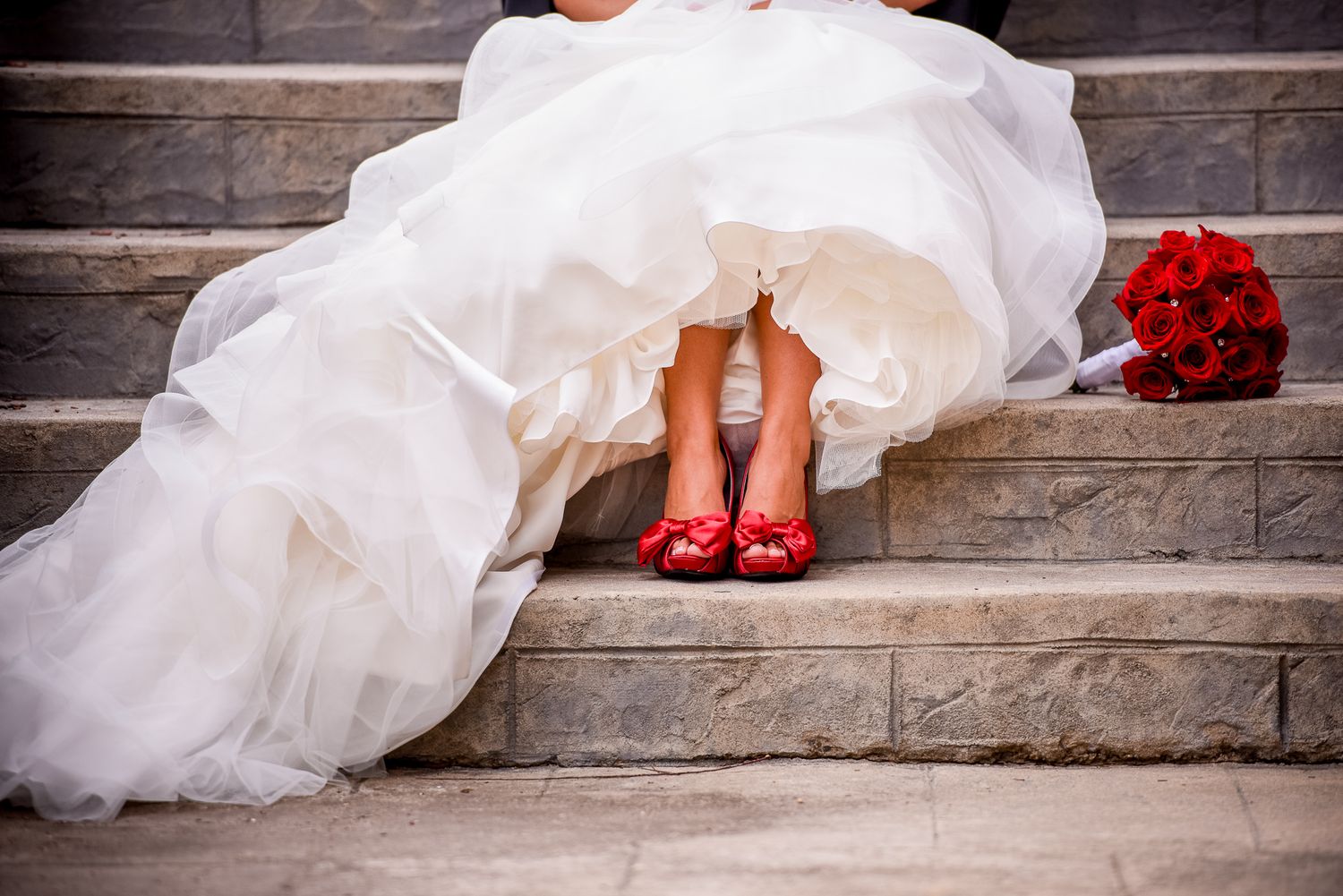 Artistic close-up of white wedding dress and red shoes on stone steps.