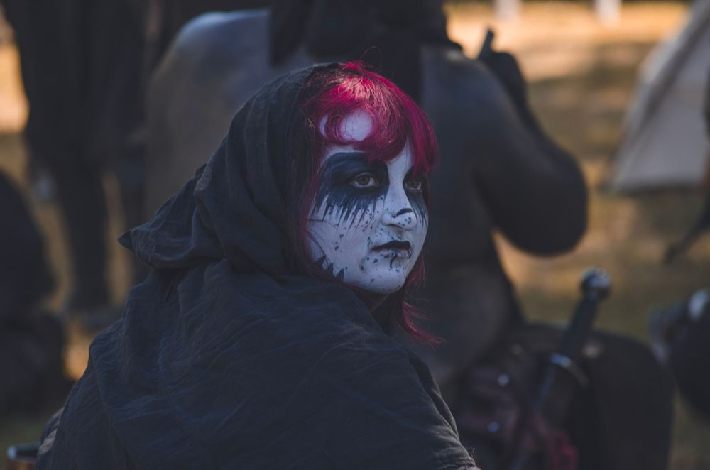Figure wearing gothic makeup and dark clothing with red hair in dramatic lighting.