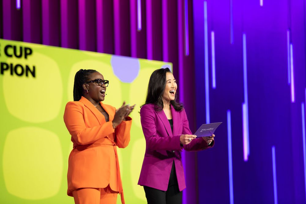 Two presenters in vibrant outfits speaking on stage with yellow backdrop and purple lighting.