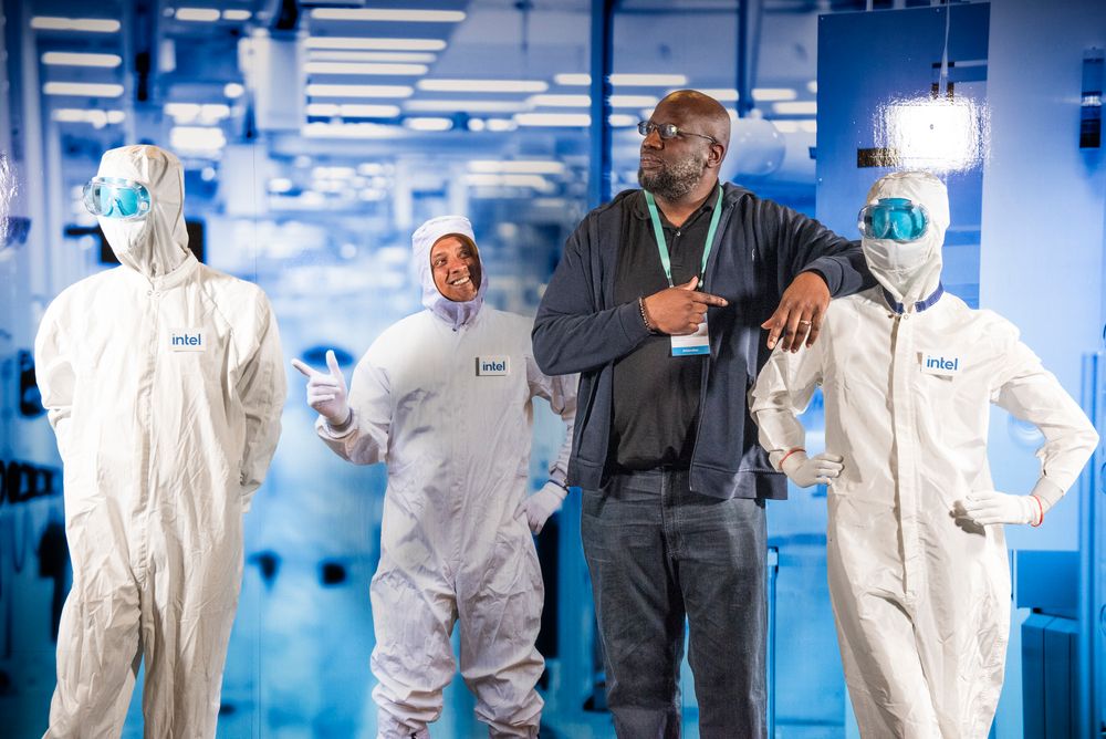 A tall figure in dark clothing stands among three people wearing white protective suits in a laboratory setting.