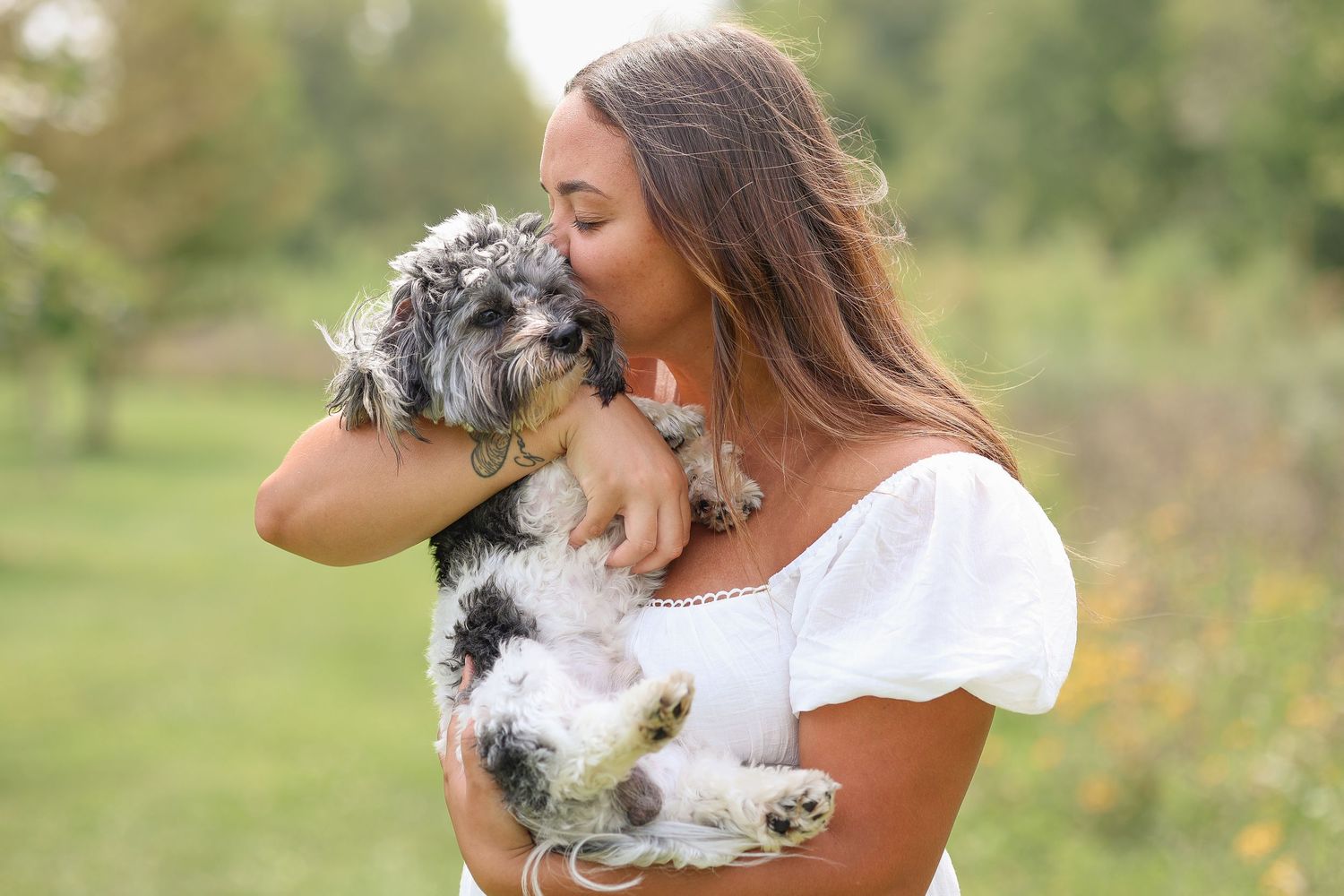 A small fluffy dog being held and kissed in a sunny outdoor setting with greenery in the background.