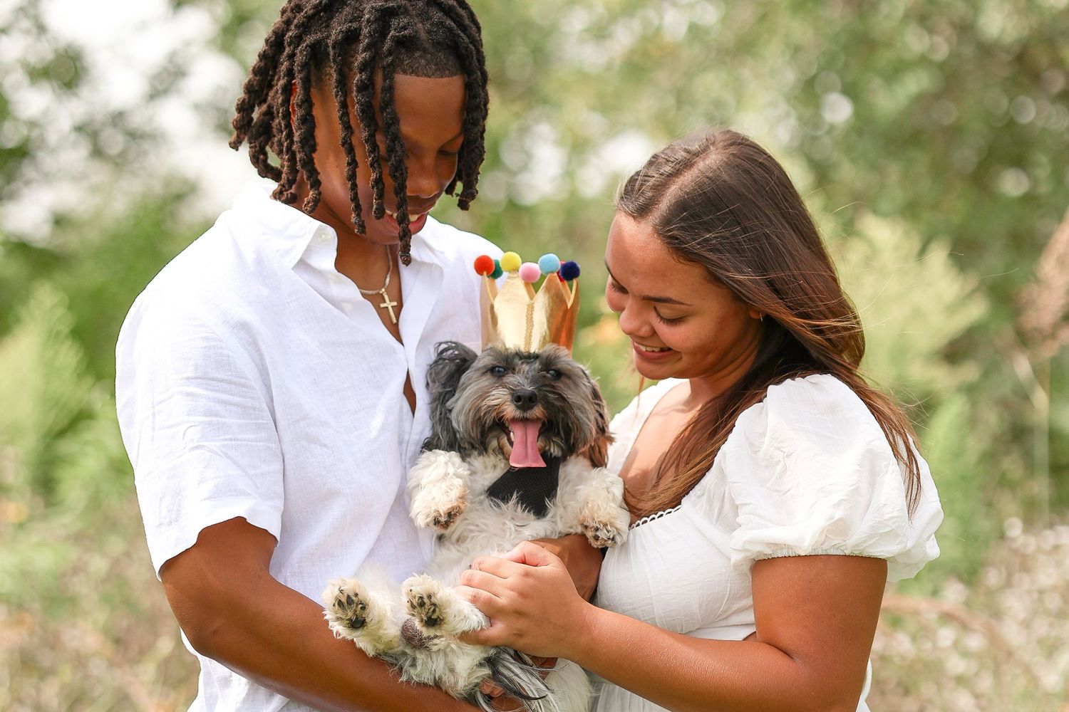 A couple shares a tender moment with their small dog in a white dress and shirt outdoors.
