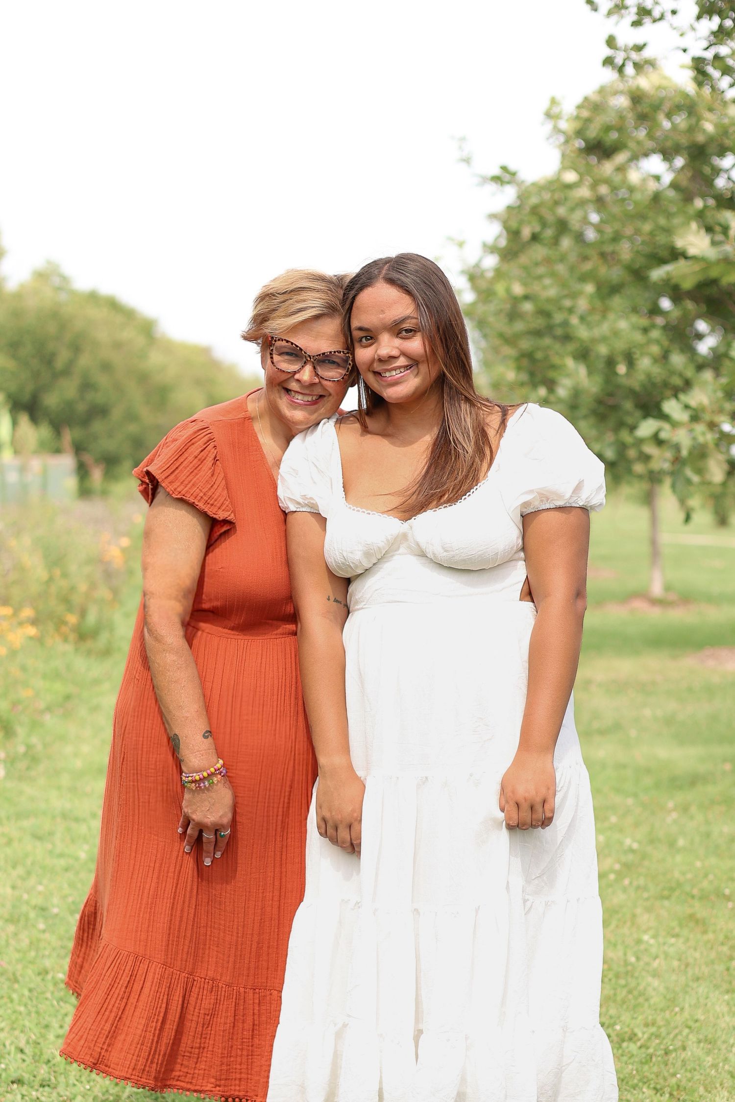 Two people embrace while standing together in a garden wearing white and orange dresses.
