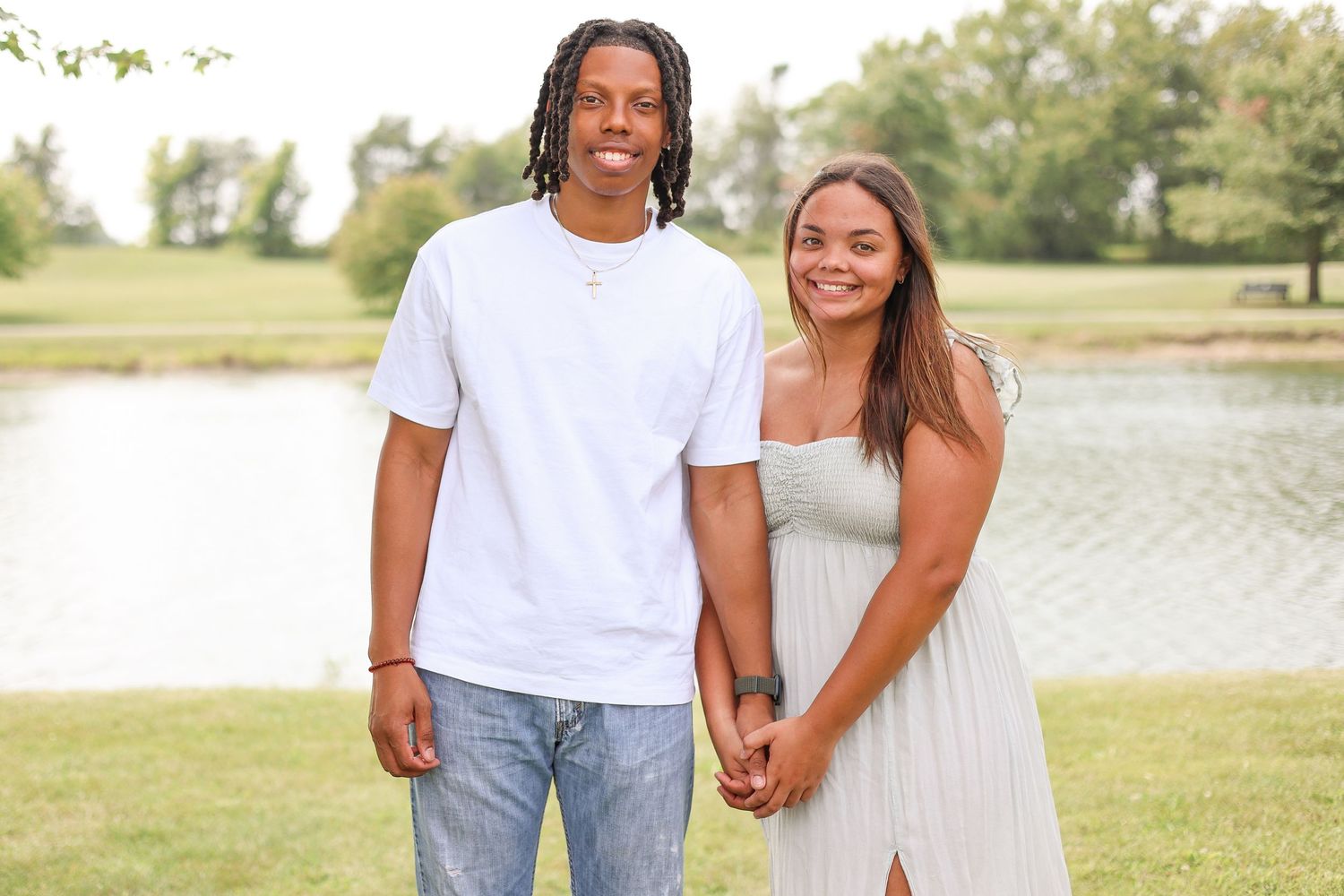 A couple stands hand in hand by a lake wearing casual white clothing on a sunny day.