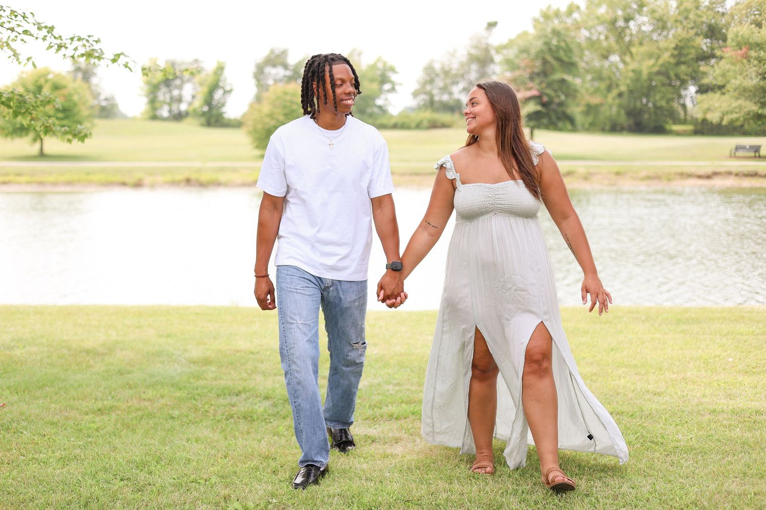 A couple walks together on grass near a lake wearing coordinated white summer outfits.