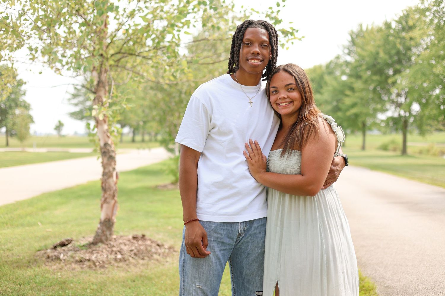 A couple embraces while standing on a tree-lined path in casual summer attire.