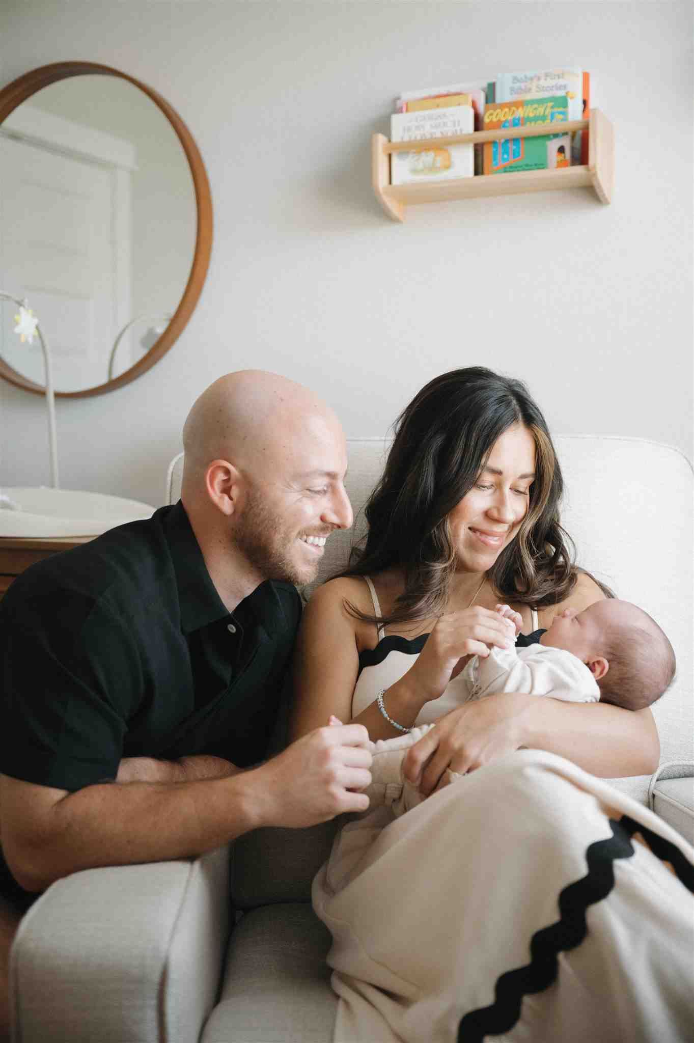 A couple sits together on a white couch with a newborn baby while a round mirror and book shelf decorate the wall.