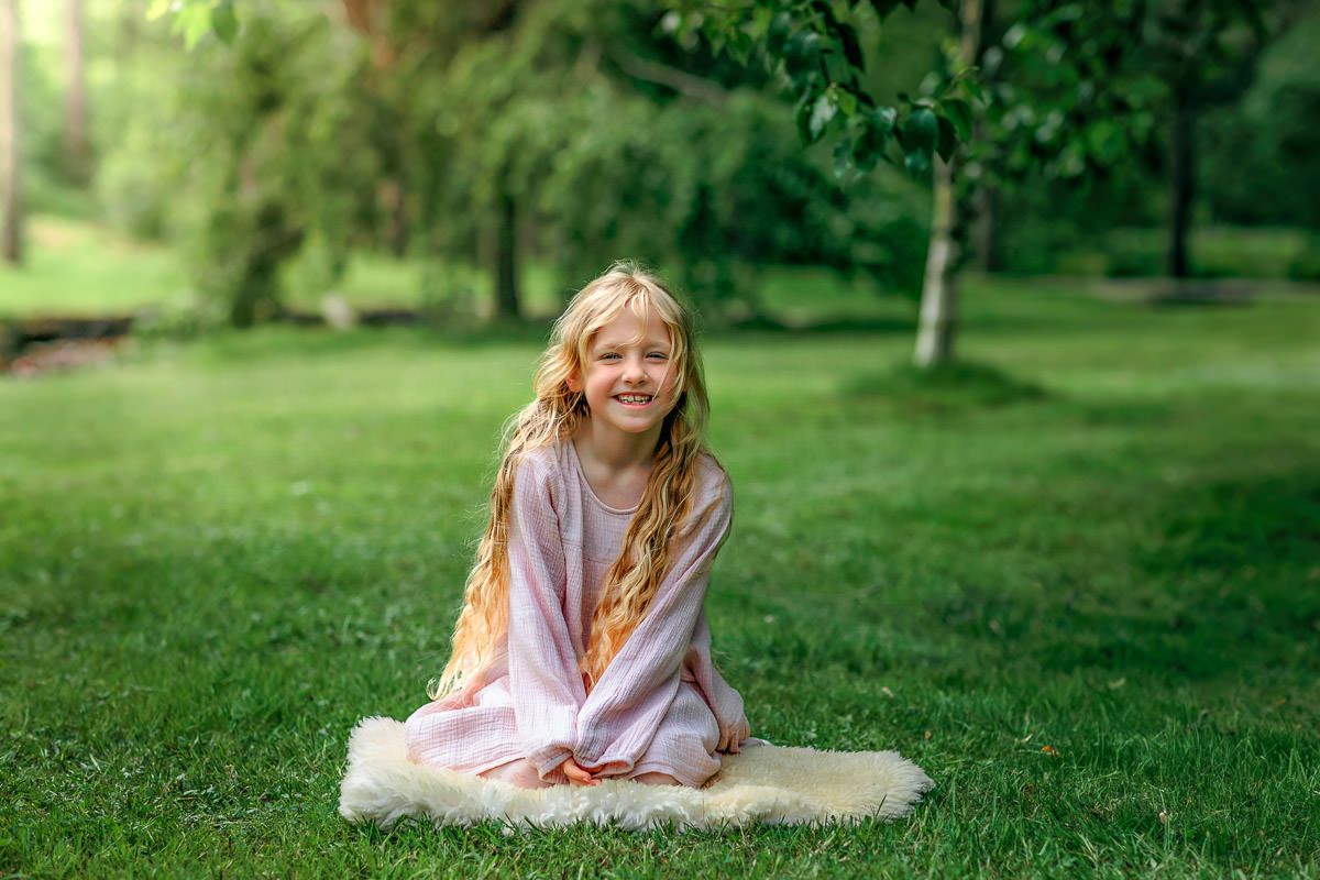 Jente på 7 år som sitter på et saueskinn og smiler til kamera. Hun er omringet av grønn plen og skog i nydelig sollys. Rogaland Arboret.