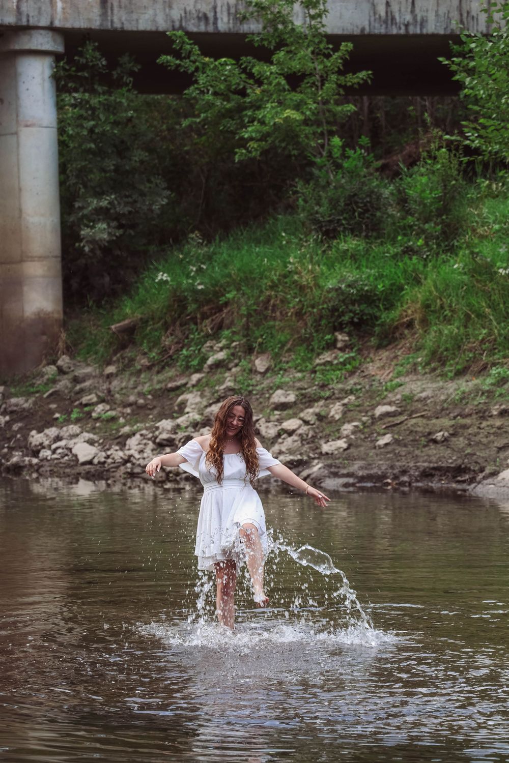 Person in white dress splashing in creek water under bridge.