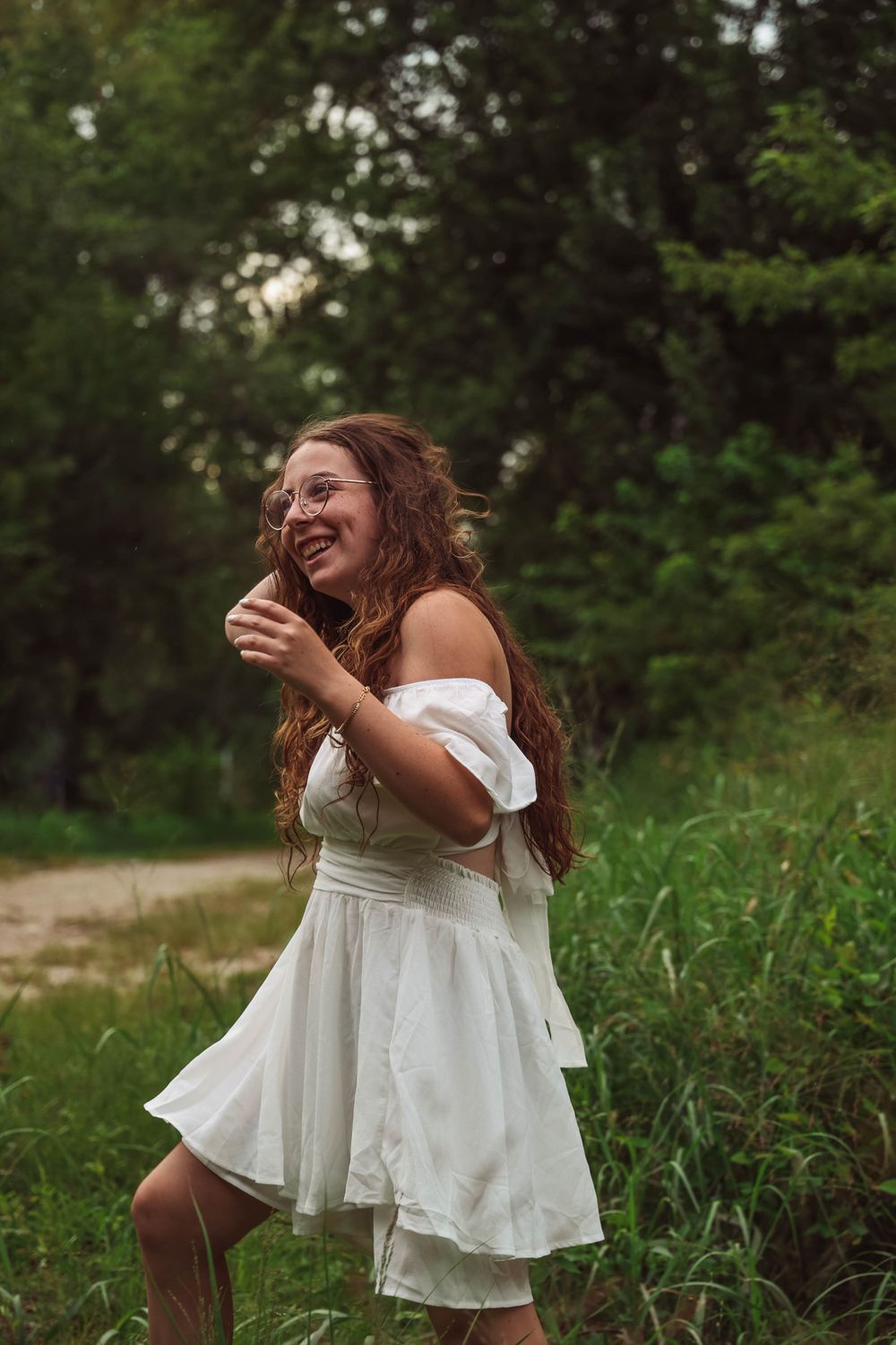 Woman in flowing white dress laughs joyfully while dancing in a green outdoor setting at sunset.