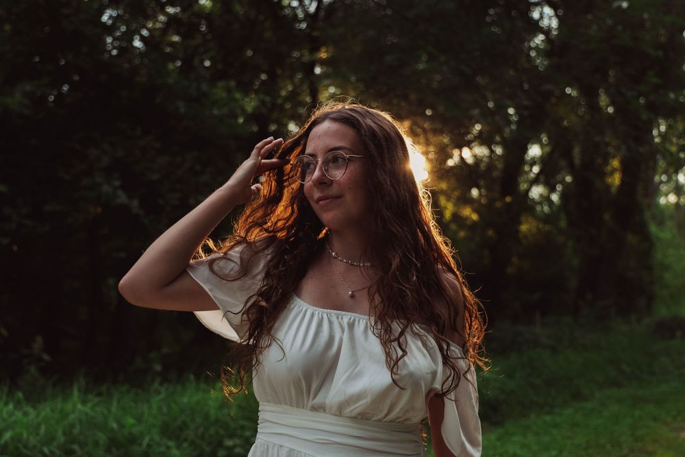 Person in white dress poses with flowing hair backlit by golden sunlight in nature.