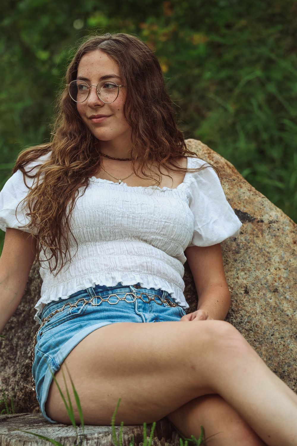 Person wearing glasses and white top relaxes on a rock in a natural setting.