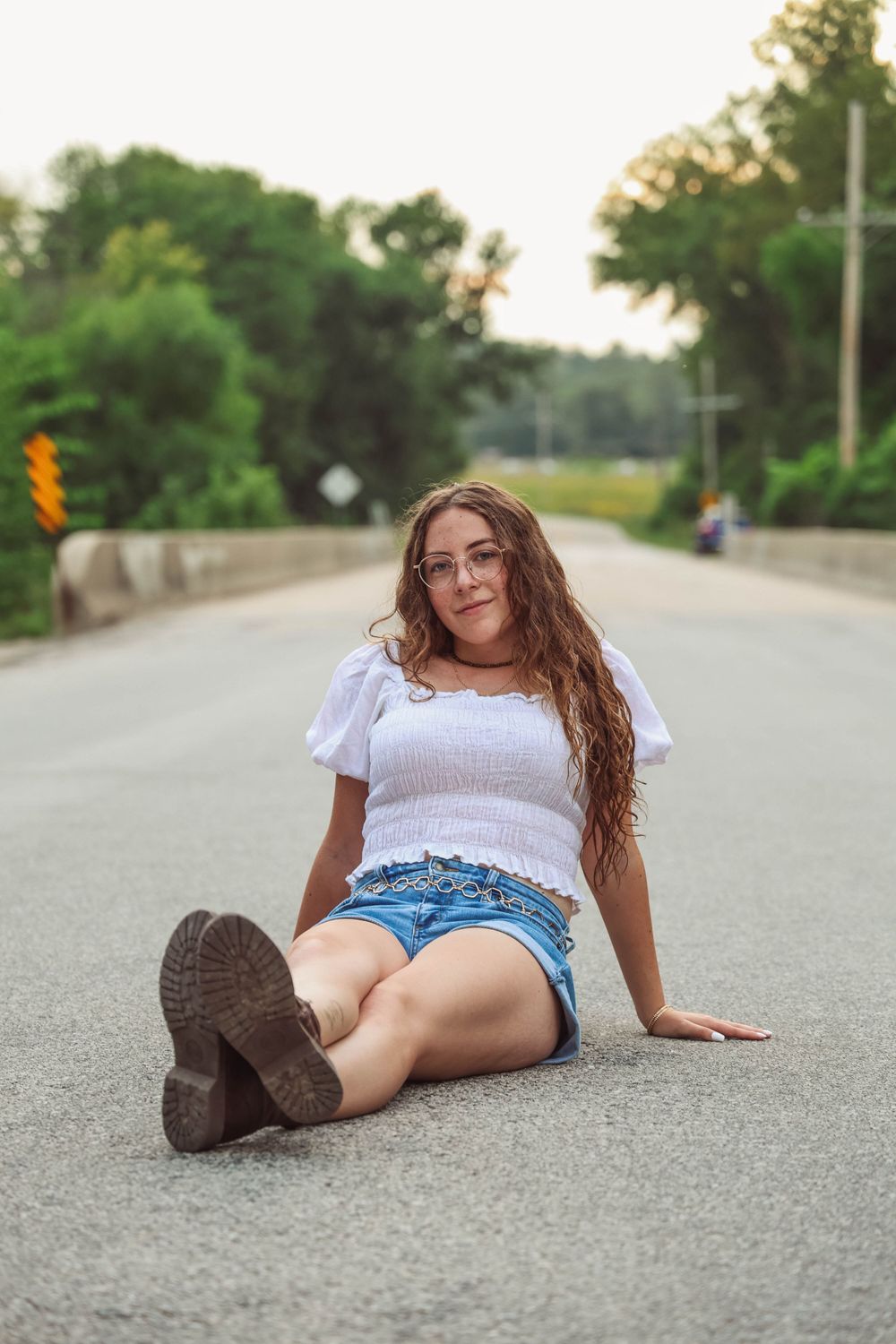 Person in white top and denim shorts sits casually on a paved road at sunset.