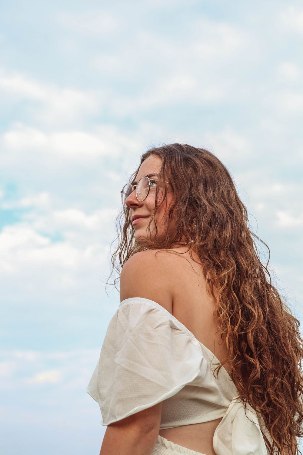 Candid profile shot against blue sky showcasing long curly hair and an off-shoulder white top.