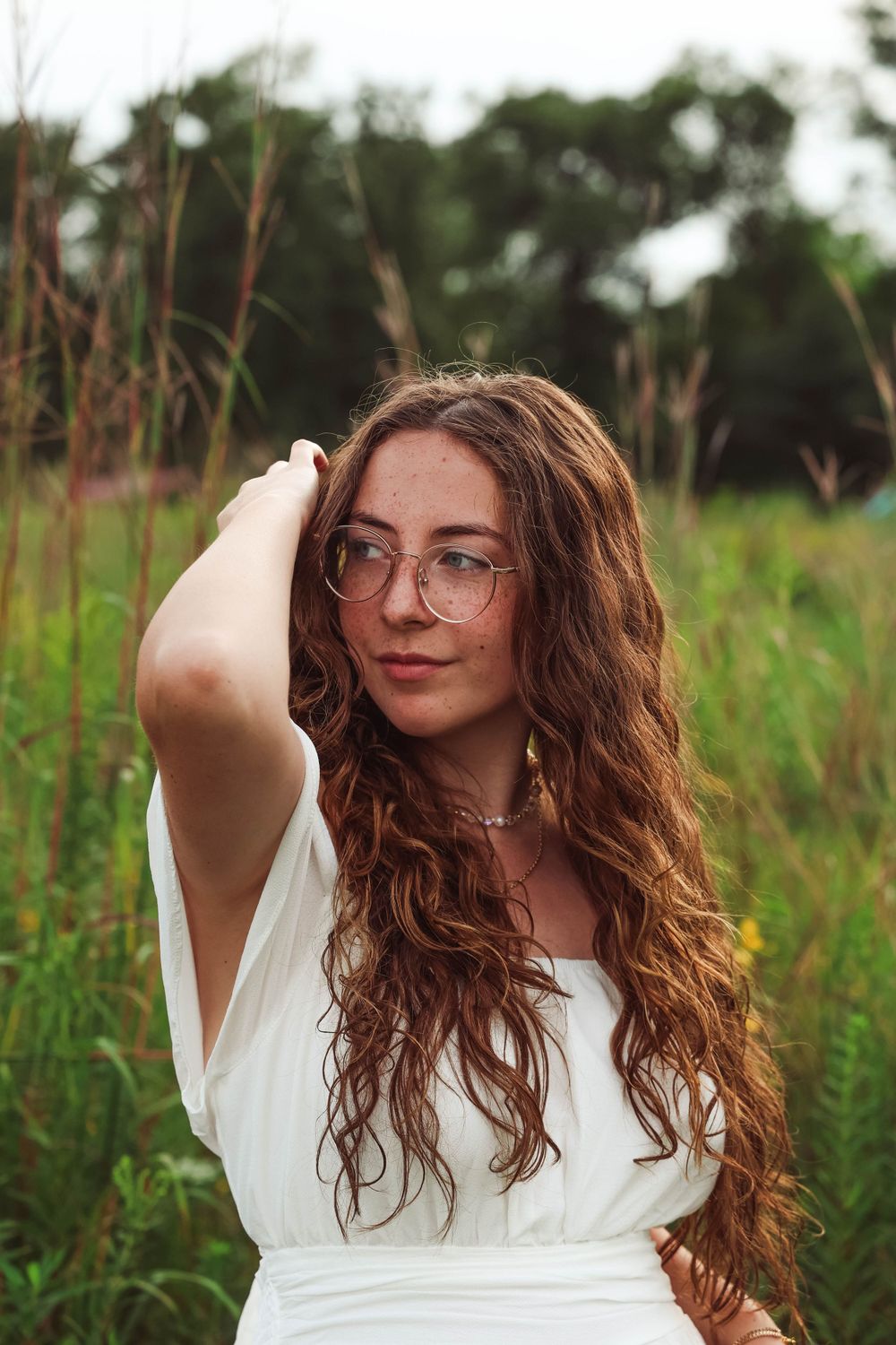 Candid outdoor portrait with long wavy hair, glasses and white dress surrounded by tall grass.