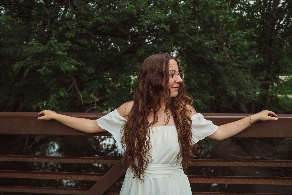 Joyful pose with arms outstretched wearing a white dress against a wooden bridge background.