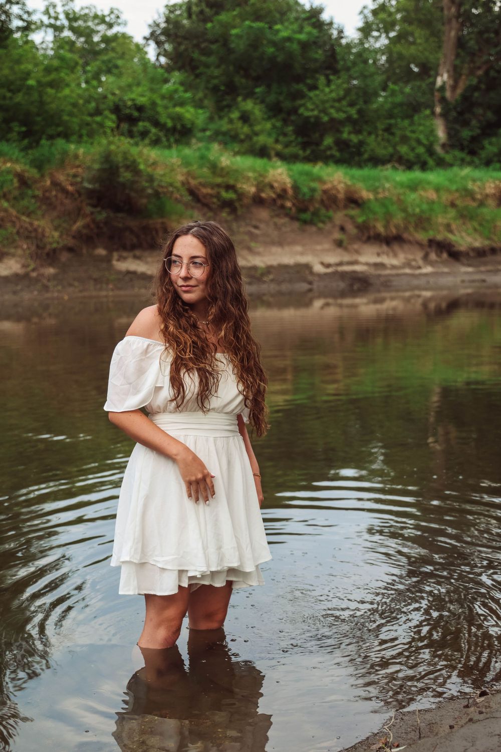 Person wearing white dress stands in shallow water with boots near muddy riverbank.