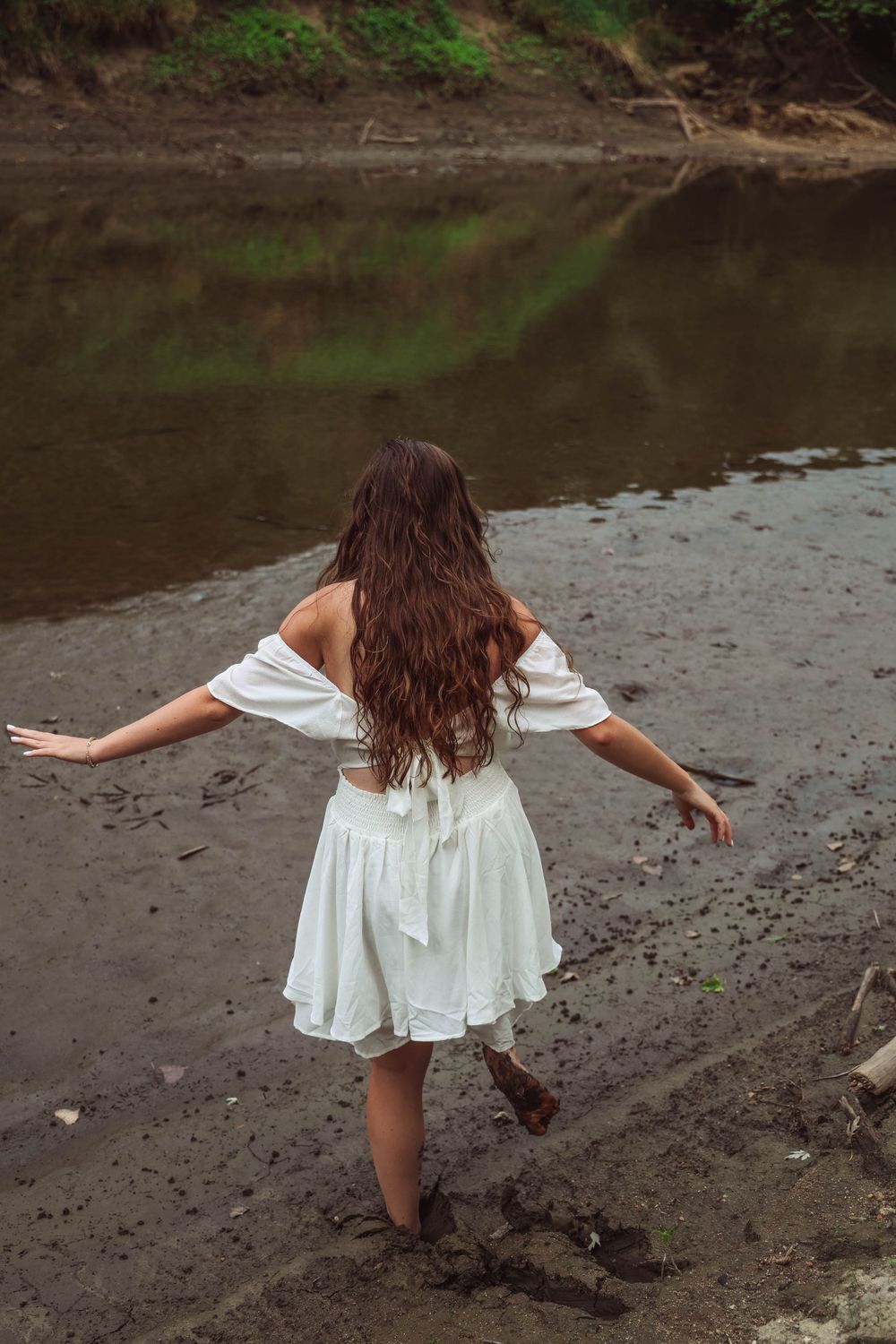 Back view of person in white dress balancing with arms outstretched by river's edge.