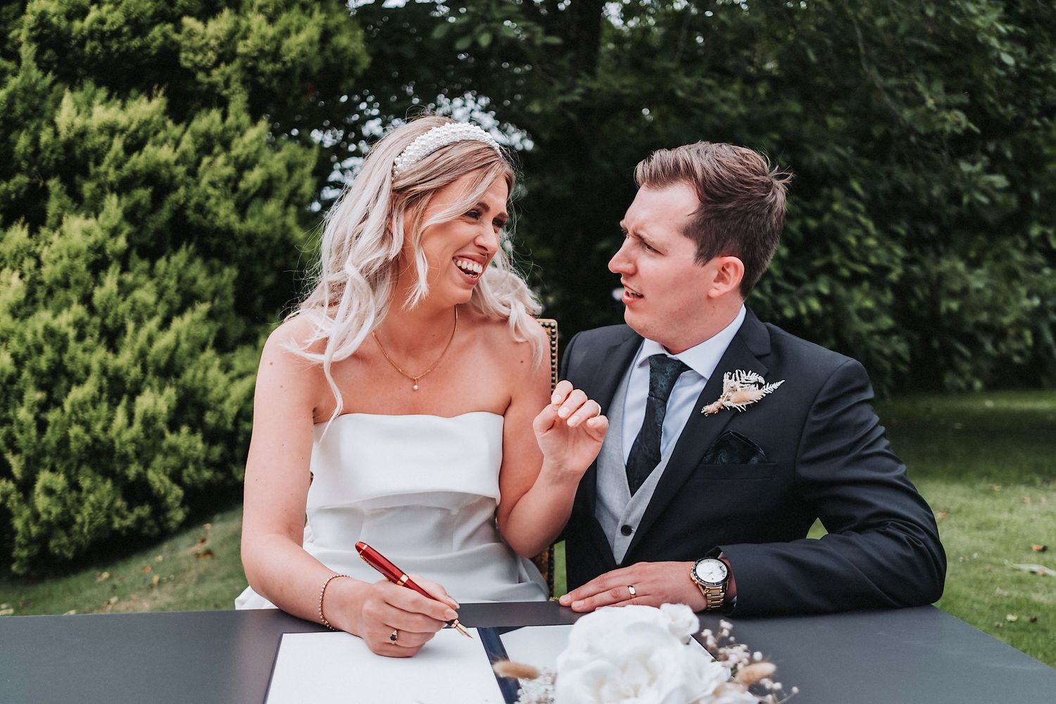 A bride and groom share a moment while signing documents at an outdoor table.