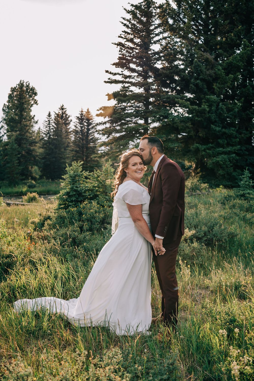 Groom kisses bride tenderly while the bride looks directly at the camera in the Teton Mountains