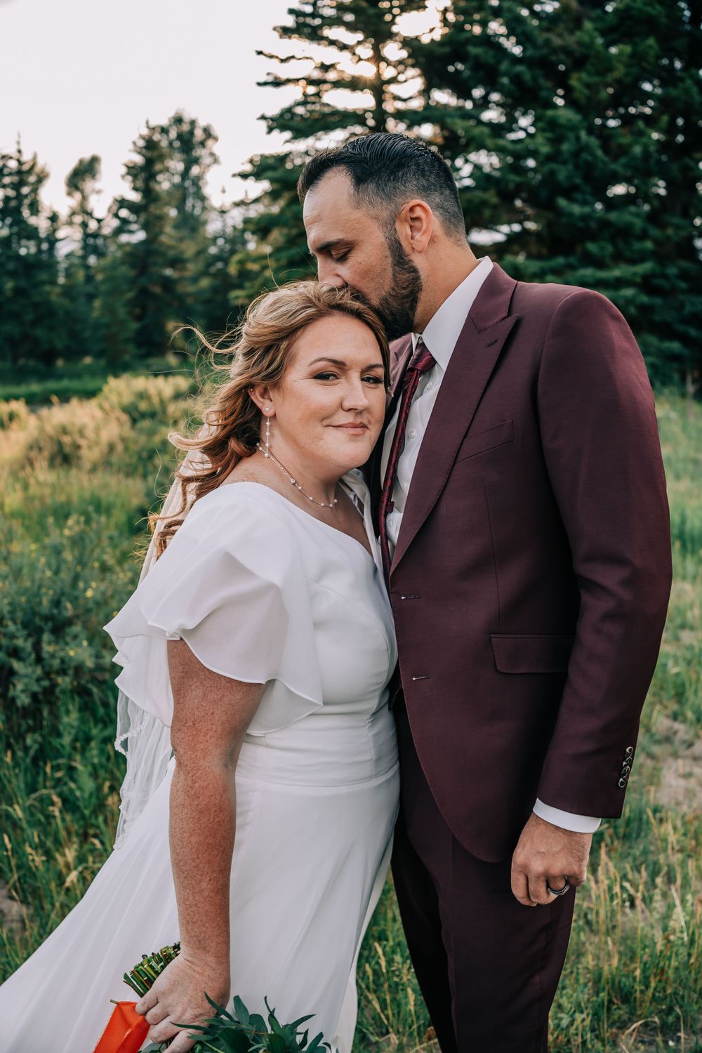 Groom tenderly kissing the top of bride's head with mountain trees as a backdrop