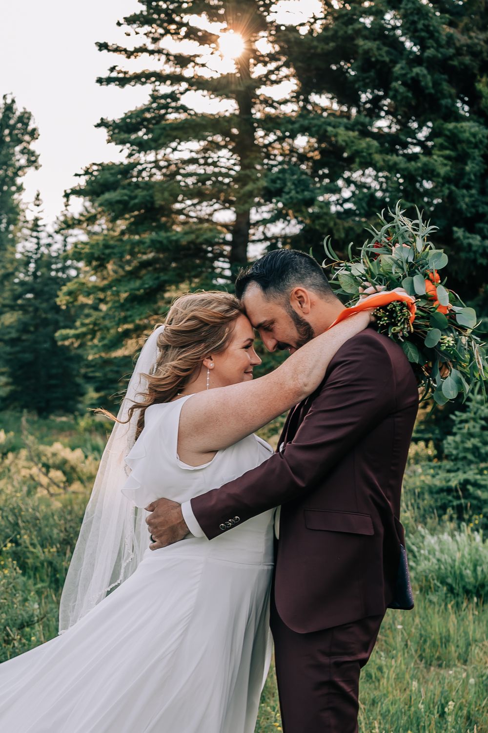 Bride and groom in a loving embrace with the sun peaking through the pine trees in the background