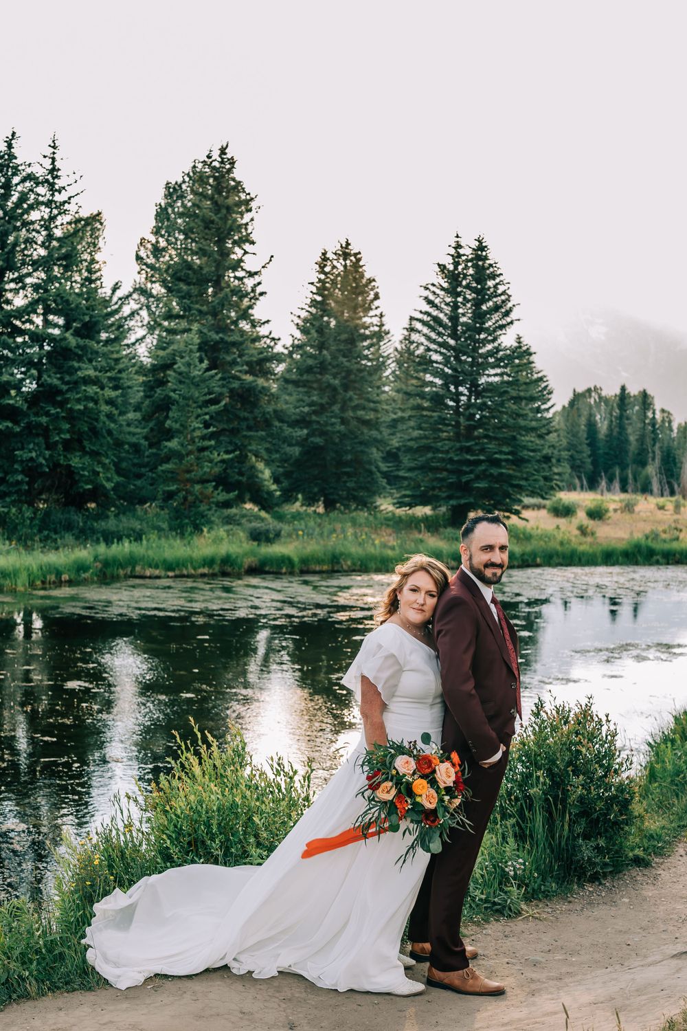 Bride hugs groom from behind standing on a Teton mountain trail near serene water