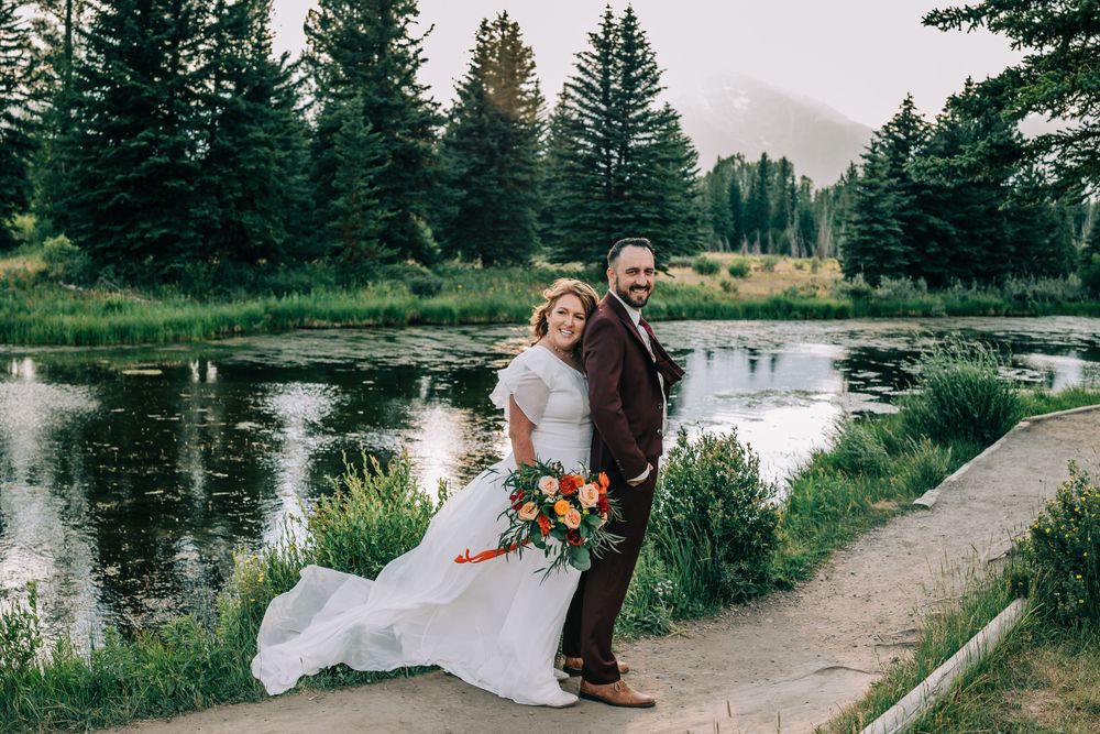 Bride hugging the groom from behind on a picturesque Teton mountain trail, with a tranquil lake and majestic mountains i