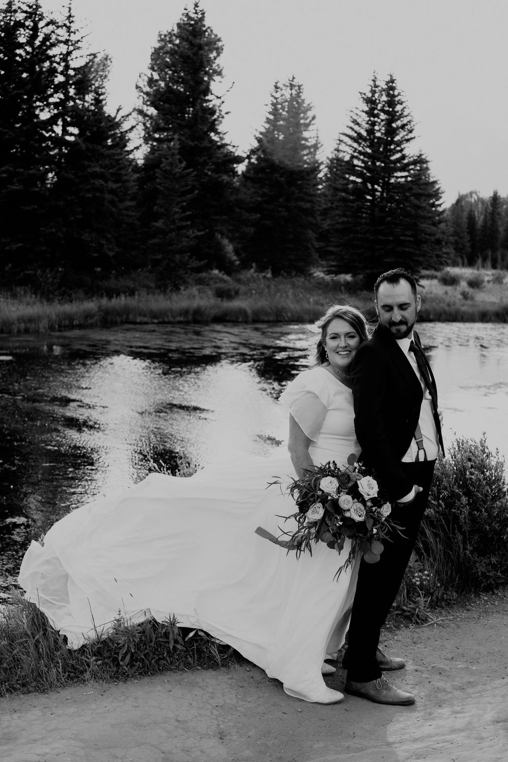 Bride wraps her arms around the groom from behind as they stand together on a Teton mountain trail, with a calm lake
