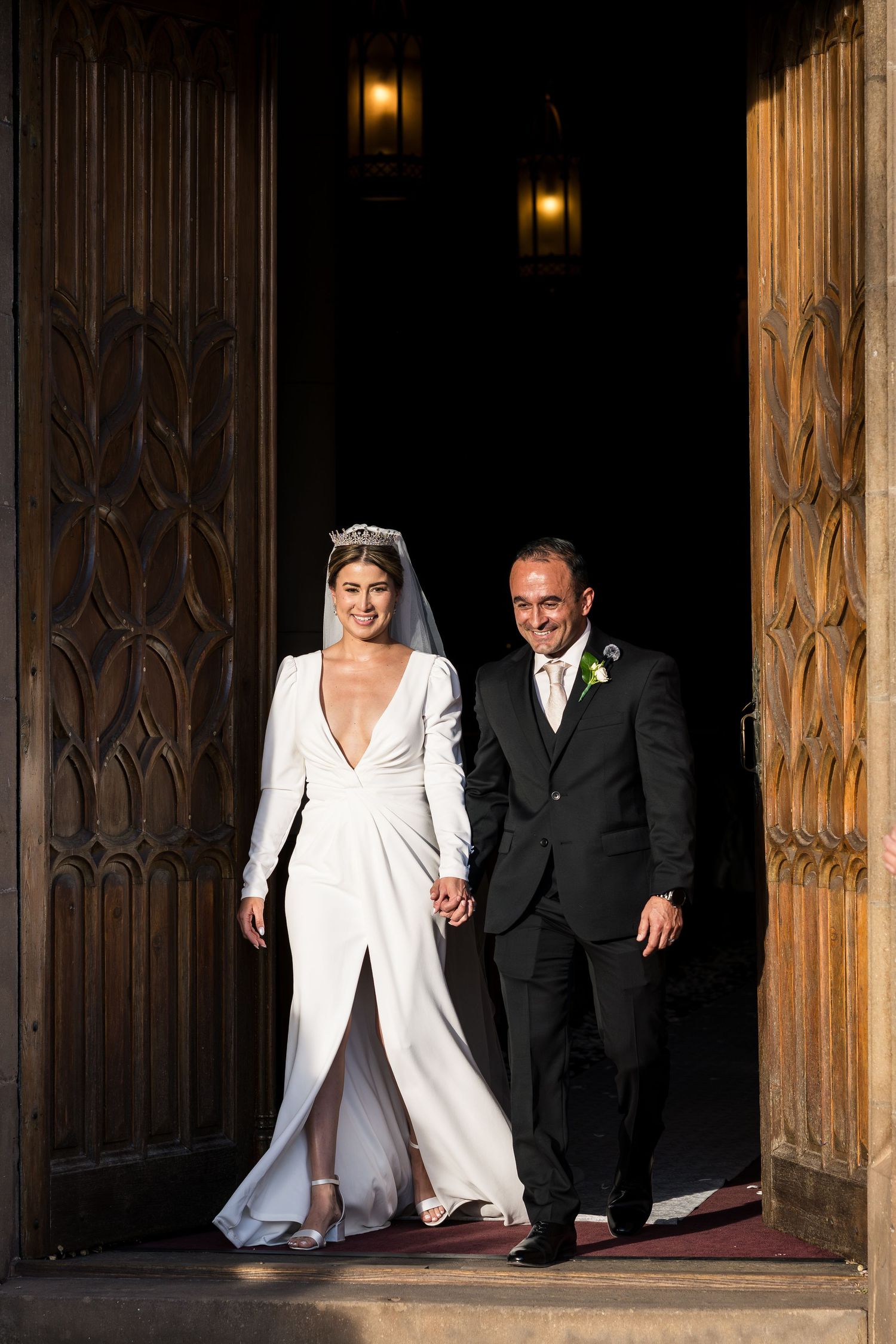 A couple emerges from ornate wooden church doors in elegant wedding attire.