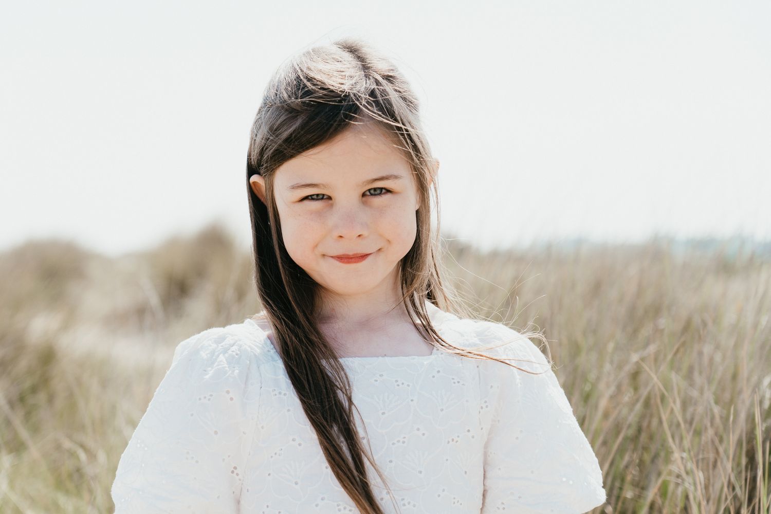 Young person in white sweater smiling against grassy coastal background.