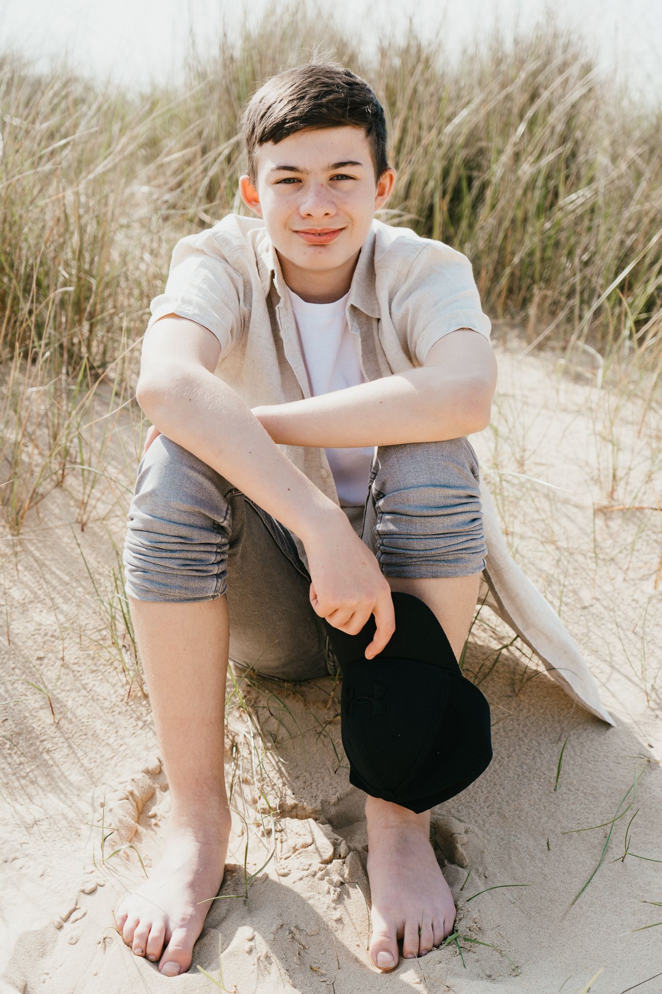 Young person sitting barefoot on sandy dunes wearing casual summer clothes.