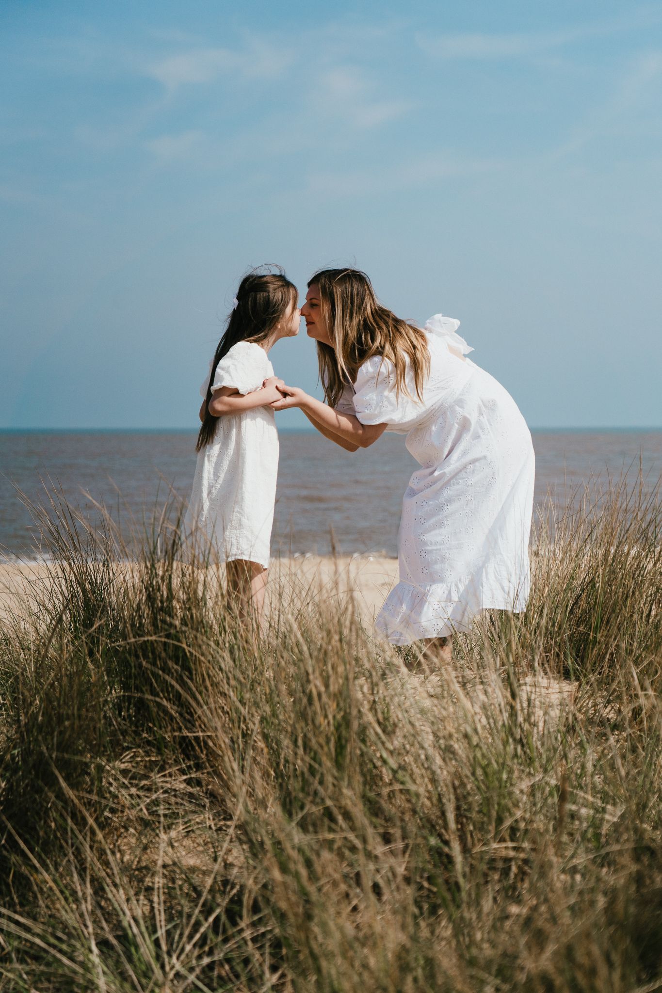 Mother and daughter in white dresses sharing tender moment by coastal grasses.
