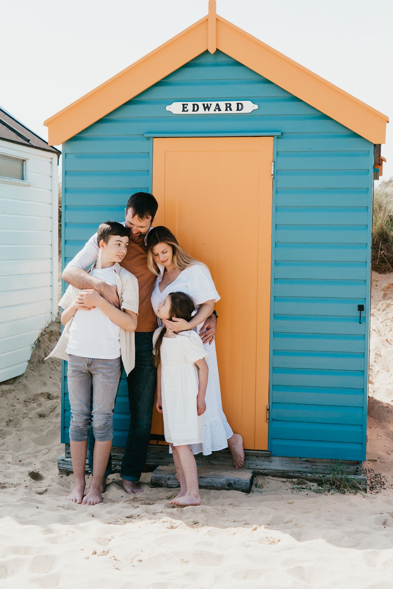 A family poses together in front of a bright turquoise beach hut with an orange roof and yellow door on a sandy beach.