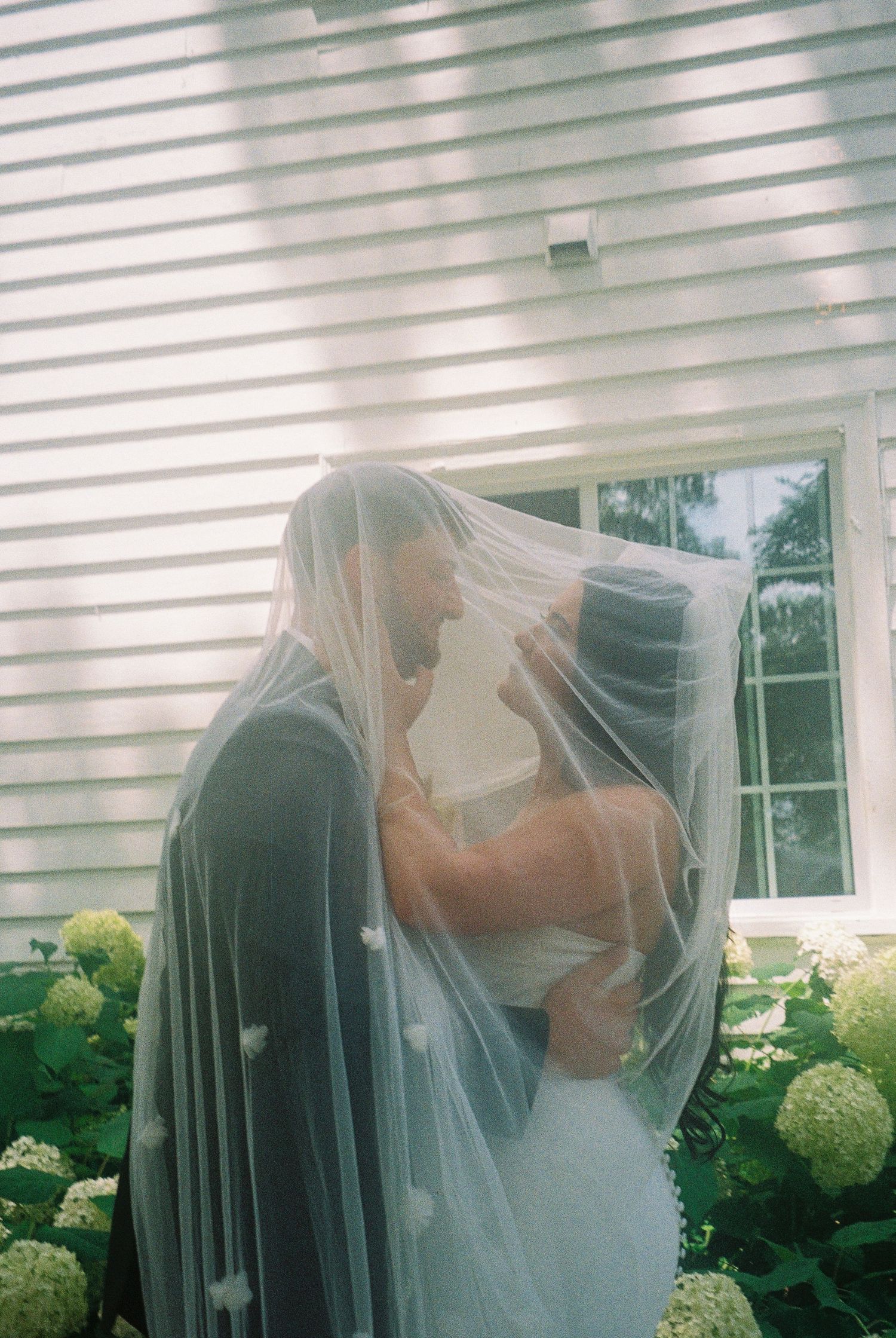 A tender moment shared under a delicate wedding veil beside a white house with blooming hydrangeas.